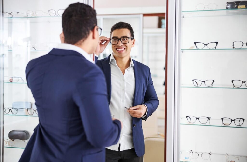 Man smiling while trying on eyeglasses in front of a mirror at an optical store, with display shelves of frames in the background.