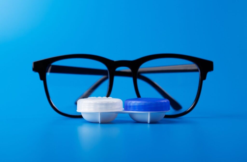 A pair of black-rimmed eyeglasses positioned behind a closed white and blue contact lens case on a solid blue background.
